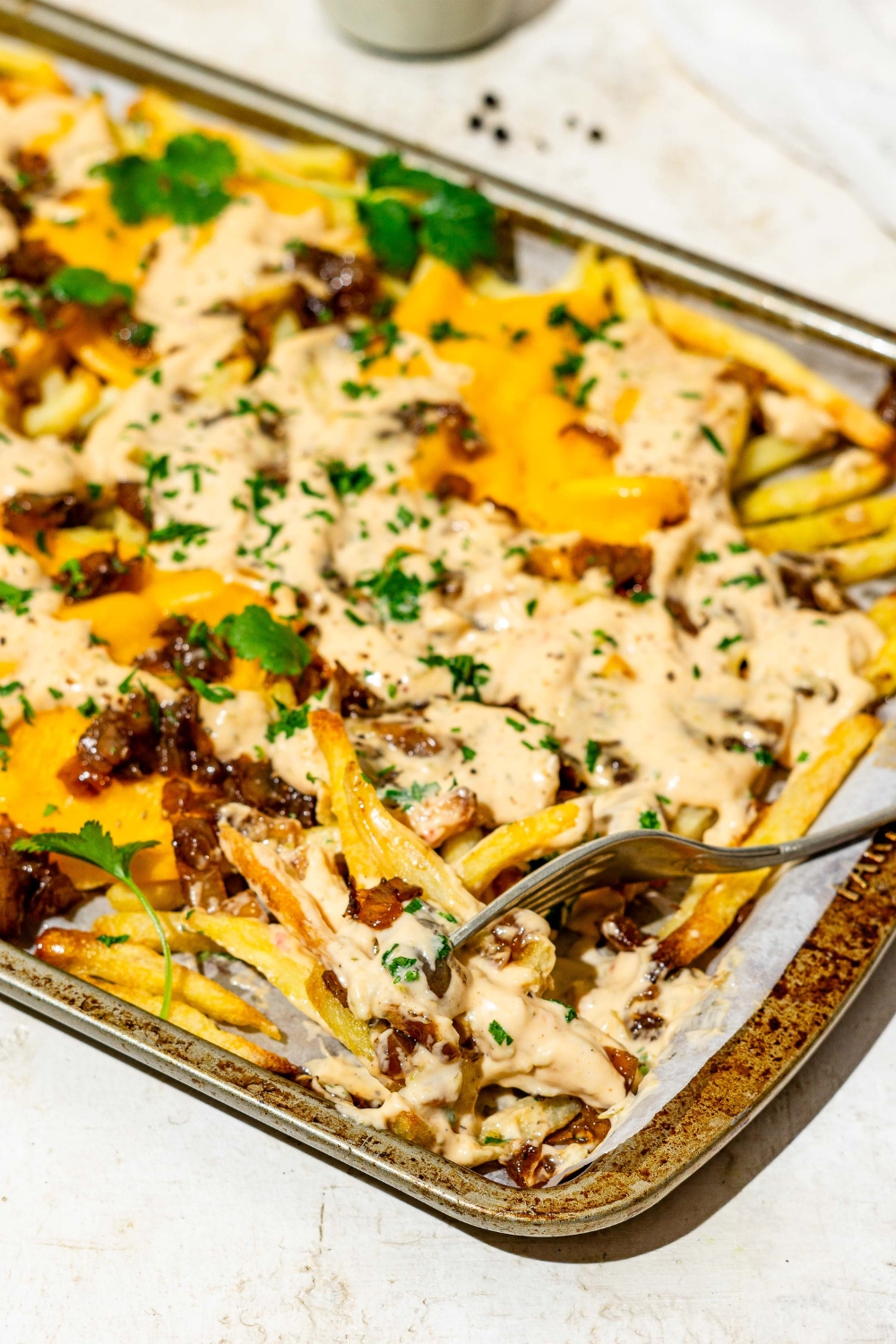 A baking sheet of In n Out animal style fries topped with melted American cheese, caramelized onions, animal sauce, and fresh parsley. A fork is taking a bite of fries. The sheet is on a tan counter.