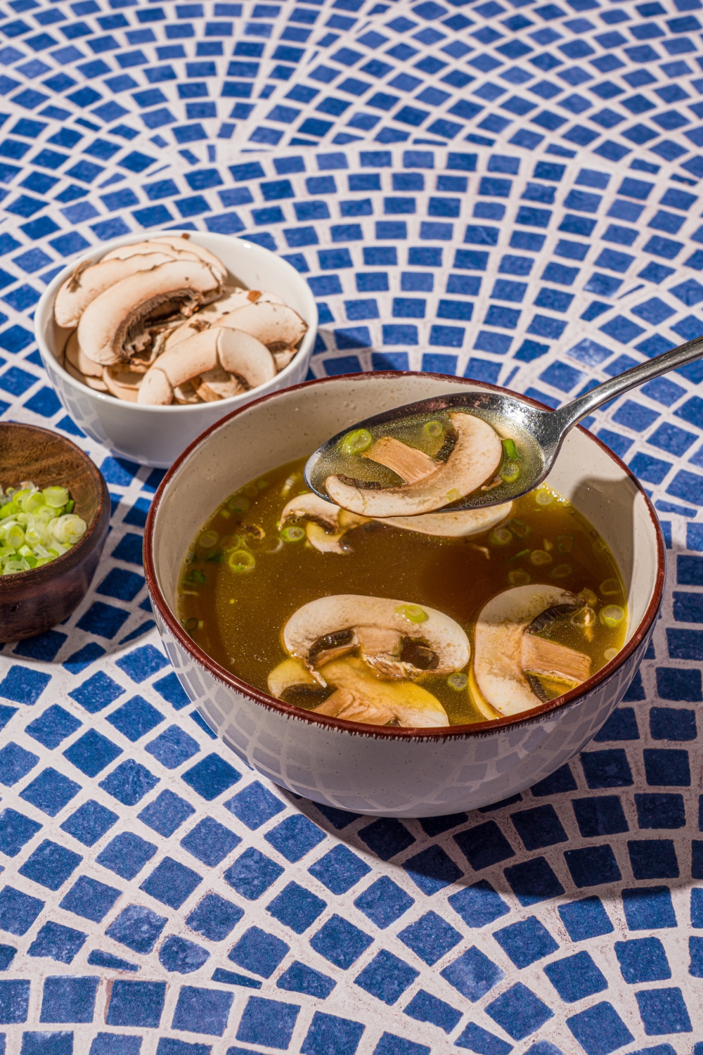 A ceramic bowl with clear soup with sliced mushrooms and green onions on a blue tiled counter with a small bowl of sliced mushrooms. There is a spoon scooping a bite of soup.