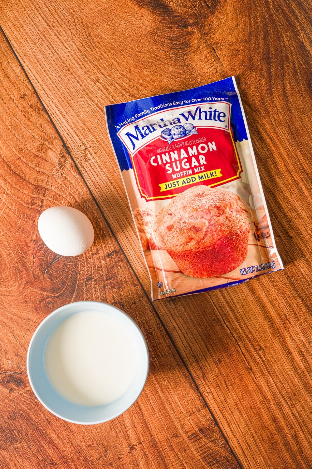 Ingredients to make muffin mix pancakes including a packet of muffin mix, an egg, and bowl of milk on a wooden counter.