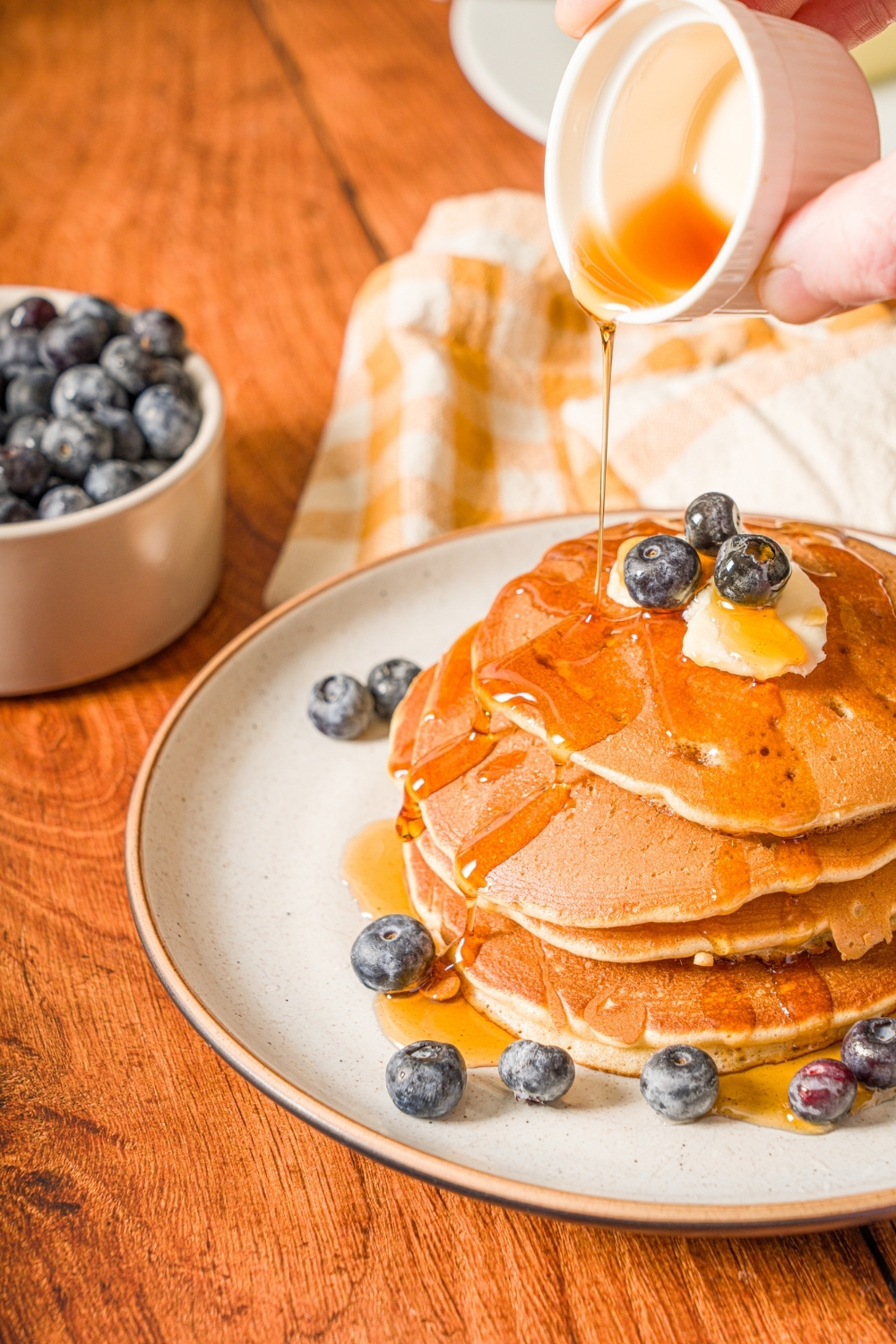 A white plate with a stack of muffin mix pancakes topped with butter and blueberries. A small cup of syrup is being poured over the pancakes. The plate is on a wooden counter with a small bowl of blueberries.