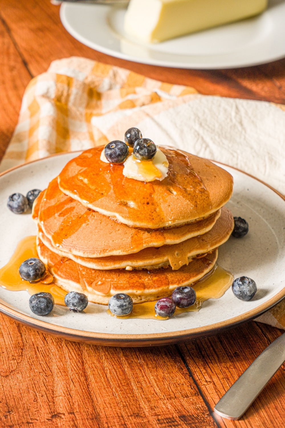 A white plate with a stack of muffin mix pancakes topped with butter and blueberries and drizzled with syrup. The plate is on a wooden counter with a small bowl of blueberries.