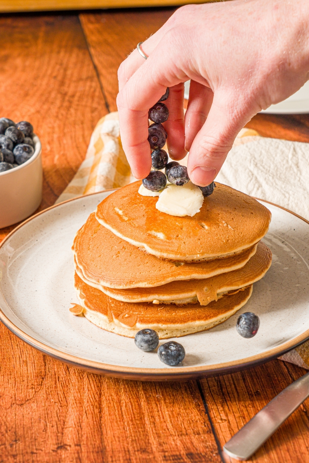 A white plate with a stack of muffin mix pancakes topped with butter. A hand is placing blueberries on the stack. The plate is on a wooden counter with a small bowl of blueberries.