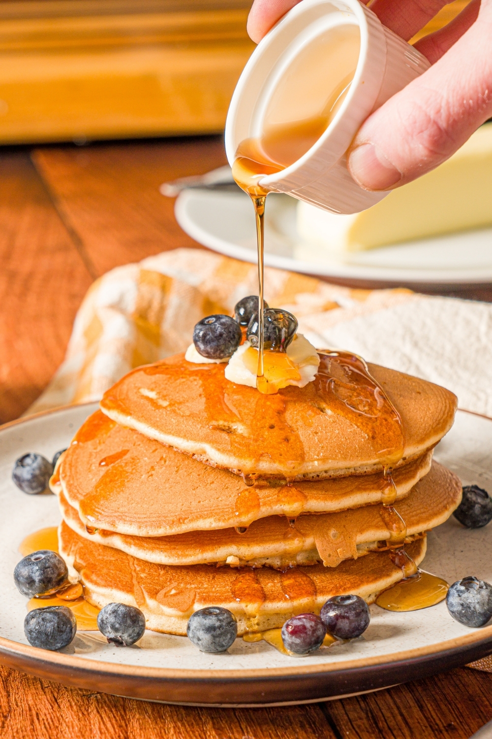 A white plate with a stack of muffin mix pancakes topped with butter and blueberries. A small cup of syrup is being poured over the pancakes. The plate is on a wooden counter with a small bowl of blueberries.