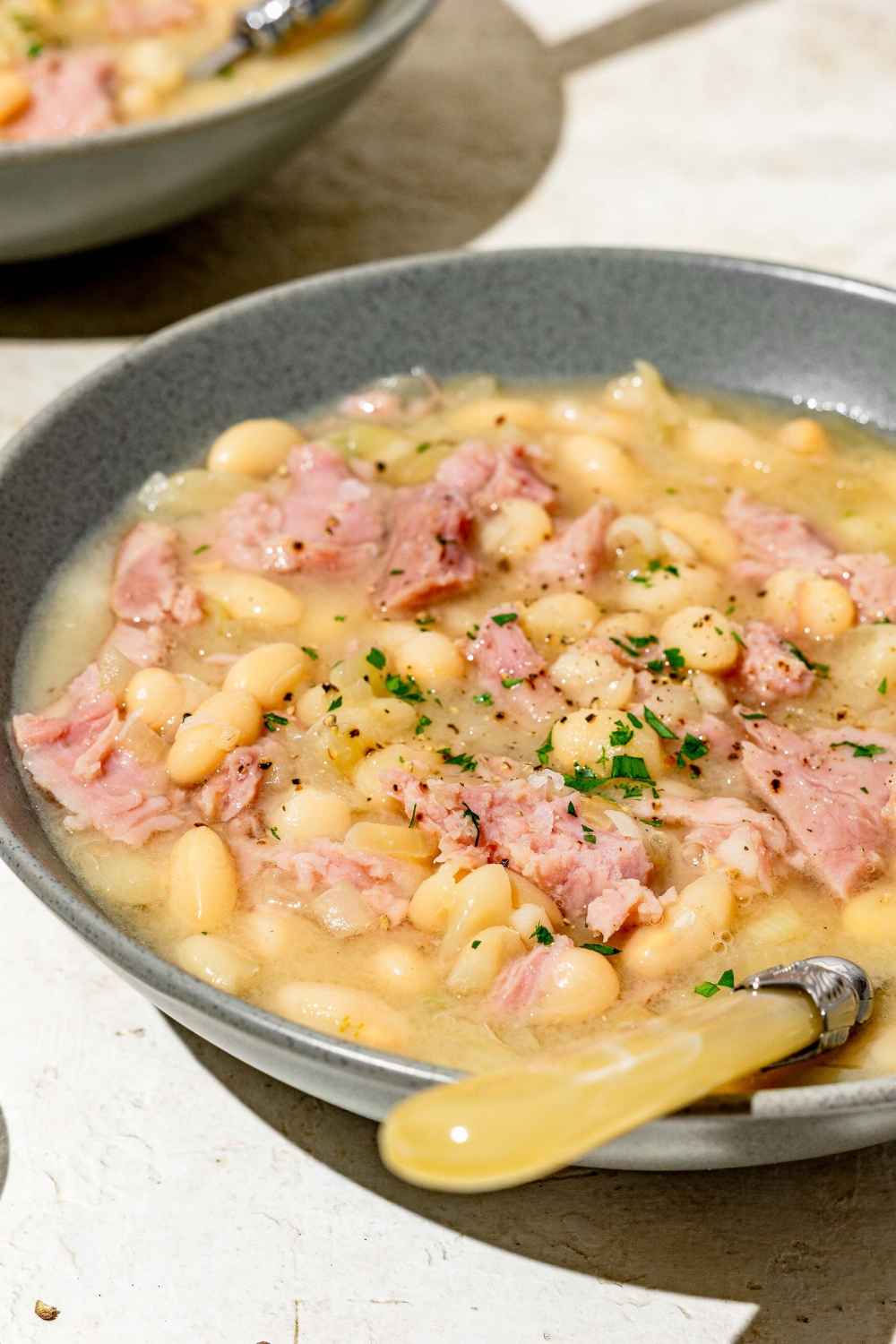 A blue bowl with senate bean soup garnished with fresh parsley. There is a spoon in the bowl. The bowl is on a white counter with an additional bowl of soup.