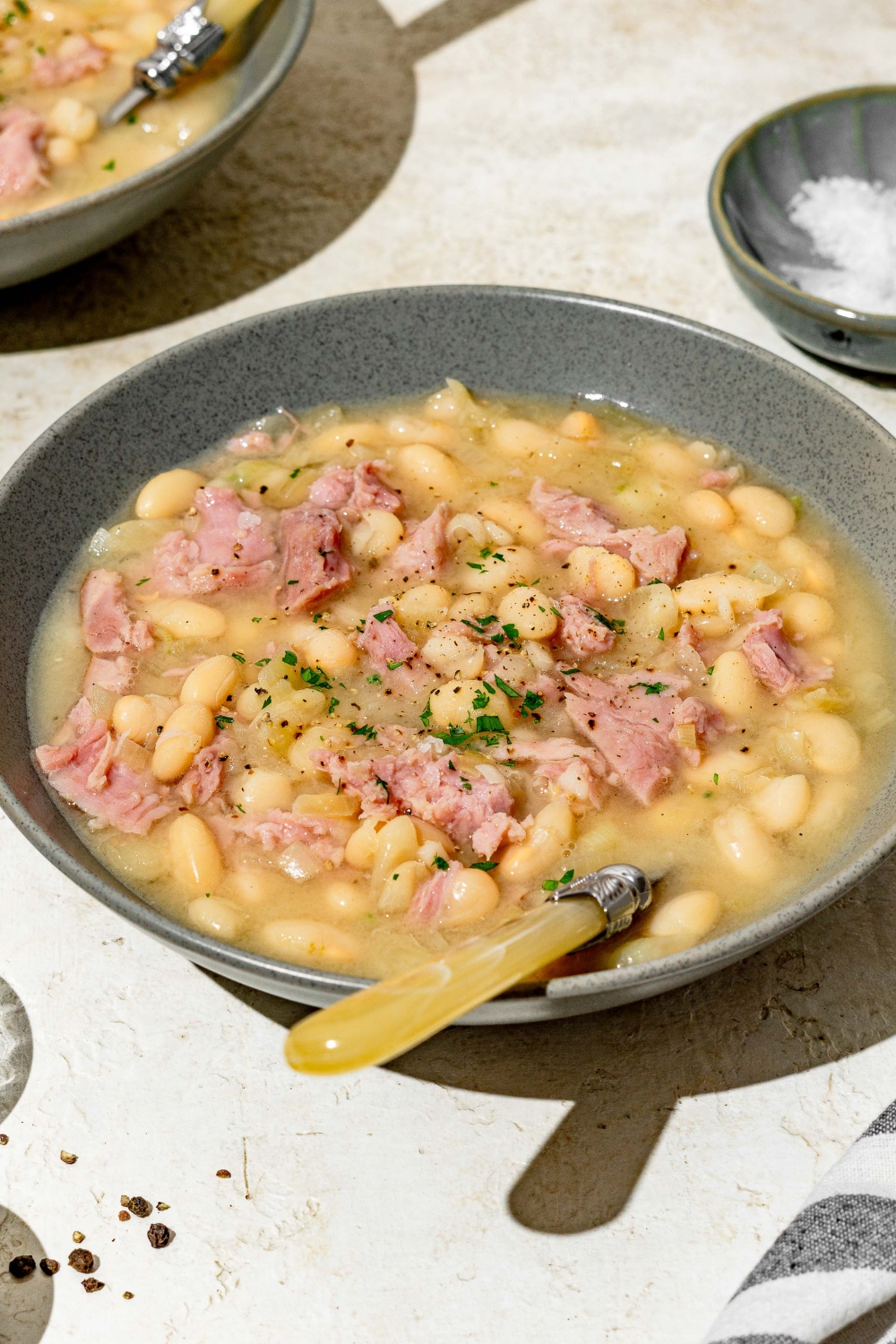 A blue bowl with senate bean soup garnished with fresh parsley. There is a spoon in the bowl. The bowl is on a white counter with an additional bowl of soup.