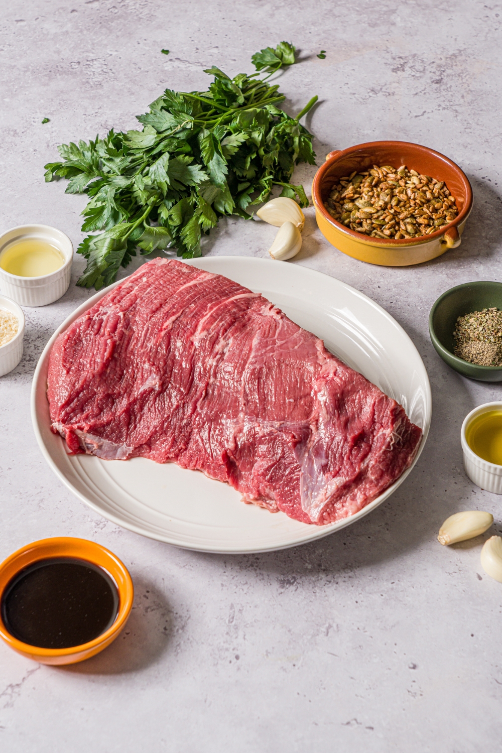 Several bowls and plates on a stone counter with ingredients to make oven baked London broil including top round steak, parsley, garlic, toasted seeds, oil, Worcestershire sauce, parmesan cheese, and garlic.