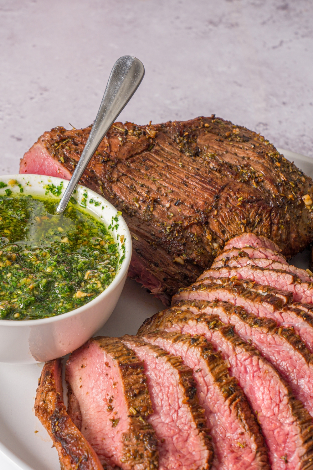 A white plate with a marinated oven baked London broil with a small bowl of pesto. Half of the steak is sliced thin on the plate. The plate is on a stone counter.