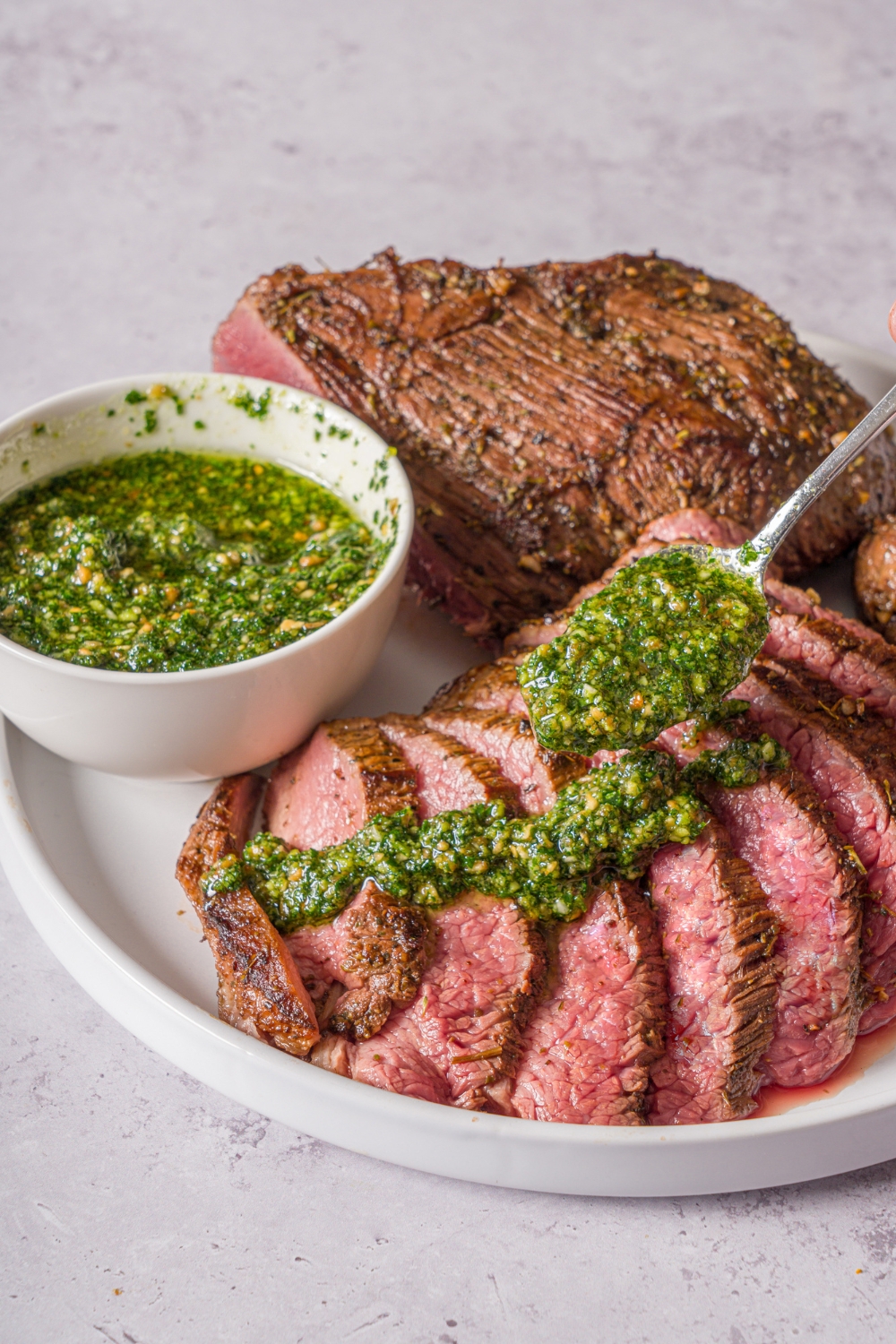 A white plate with oven baked London broil. Half of the steak is sliced thin with a spoon drizzling pesto over the slices. There is a bowl of pesto on the plate. The plate is on a stone counter.