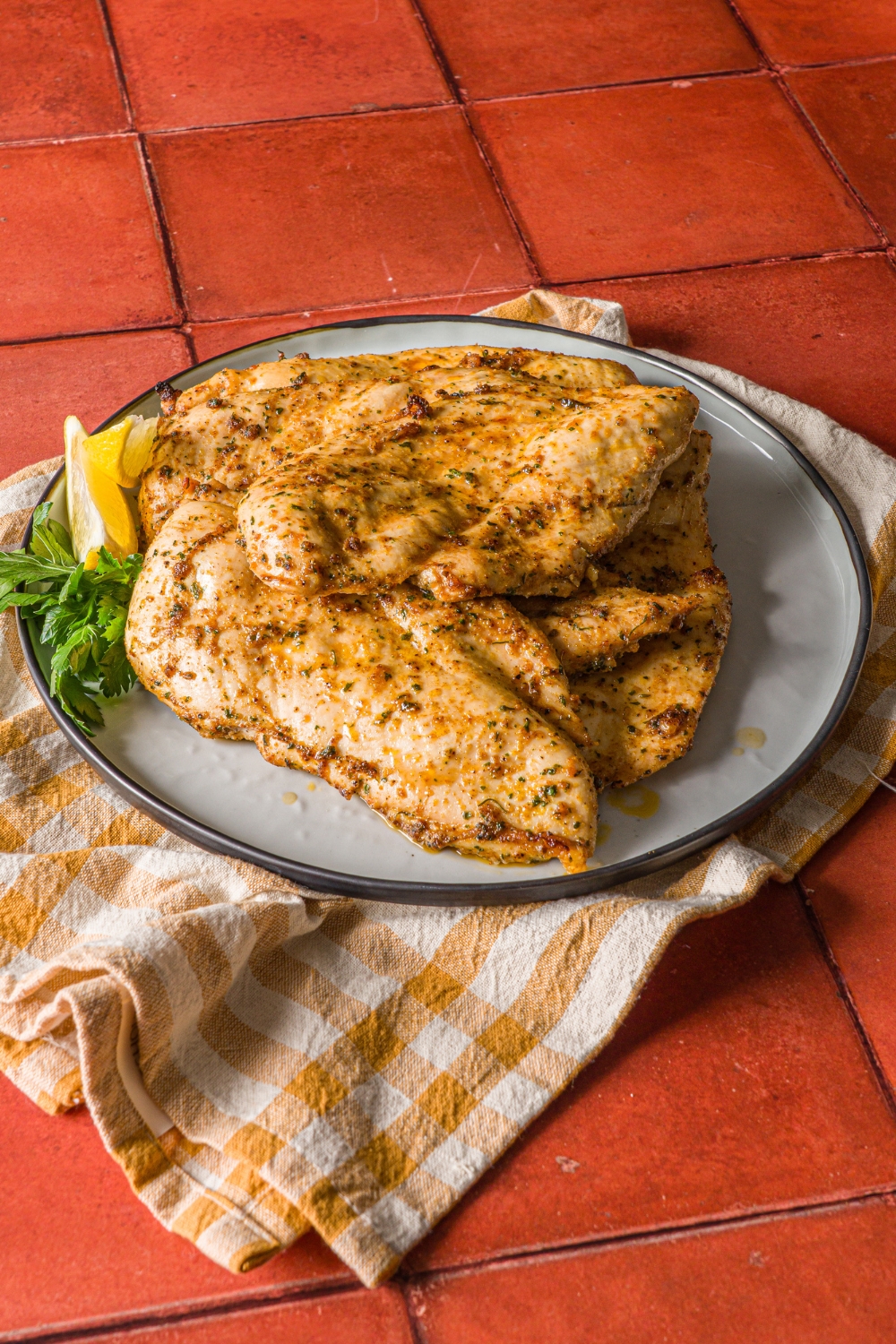 A blue plate with thin sliced oven baked chicken breasts. The chicken is cooked in seasonings and served with lemon wedges. The plate is on a yellow checkered napkin on a red tiled counter.