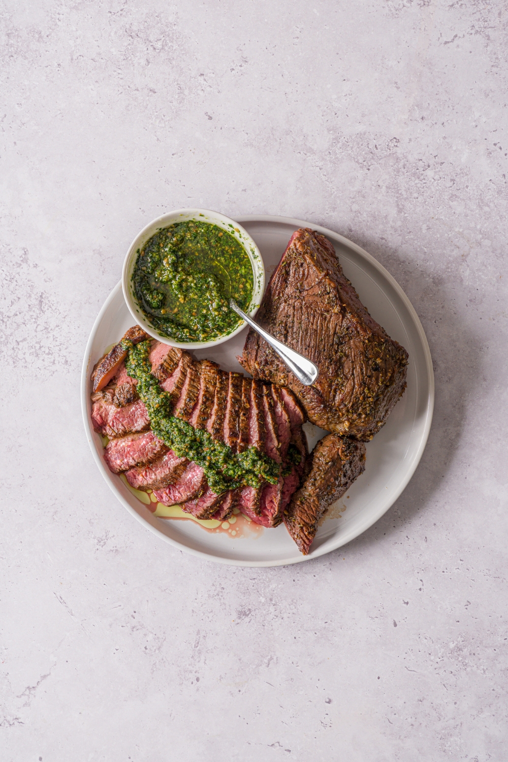 A white plate with oven baked London broil. Half of the steak is sliced thin and topped with pesto. There is a small bowl of pesto with a spoon on the plate. The plate is on a stone counter.