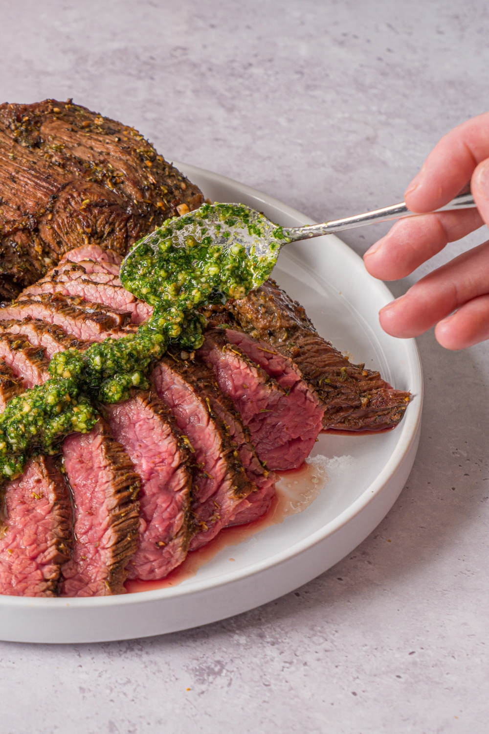A white plate with oven baked London broil. Half of the steak is sliced thin with a spoon drizzling pesto over the slices. There is a bowl of pesto on the plate. The plate is on a stone counter.