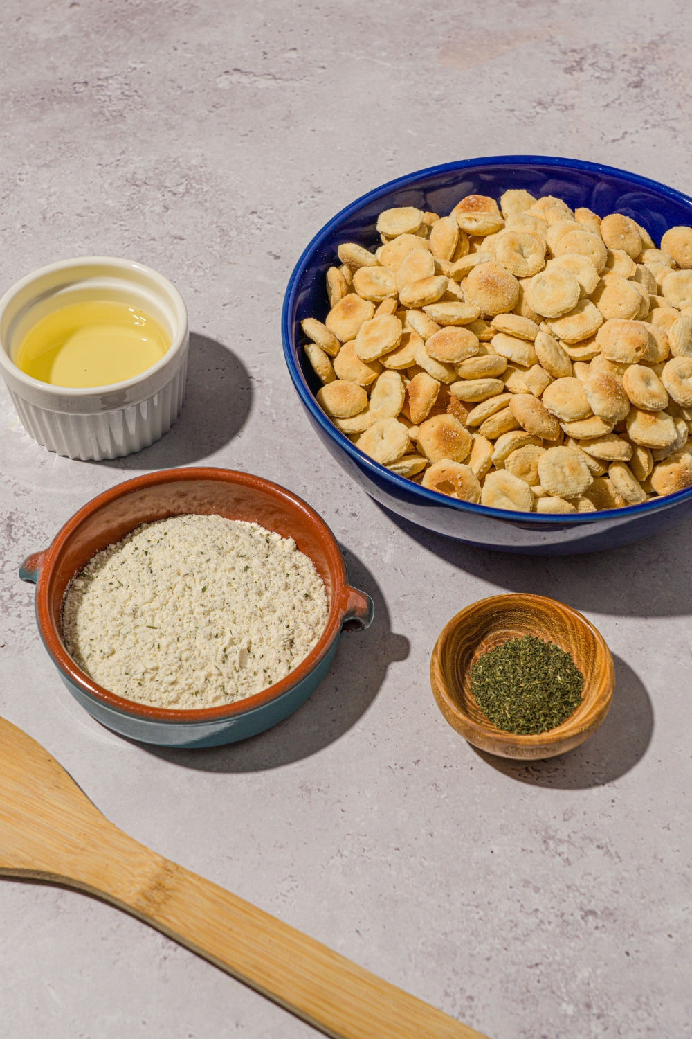 Several bowls in various sizes containing ingredients to make ranch oyster crackers including oyster crackers, oil, ranch seasoning, and dill.