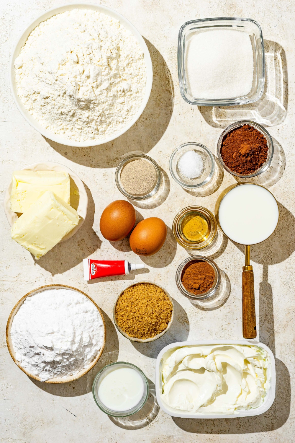 An overhead shot of several bowls in various sizes containing ingredients to make red velvet cinnamon rolls including flour, milk, sugar, eggs, butter, cream cheese, cocoa powder, red food dye, oil, and cinnamon.