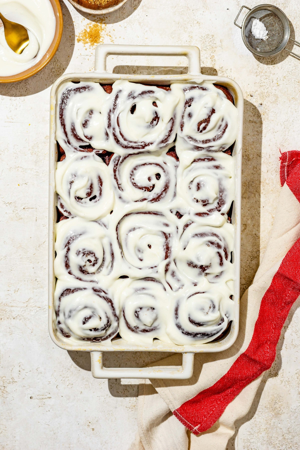 A baking dish with red velvet cinnamon rolls topped with cream cheese frosting. The dish is on a tan counter with a red striped napkin.