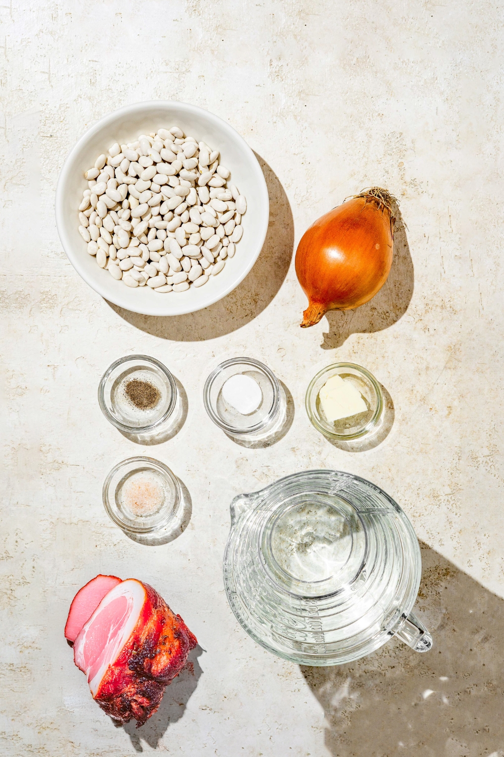 An overhead shot of several bowls in various sizes containing ingredients to make senate bean soup including navy beans, onion, water, butter, ham hock, and baking soda.