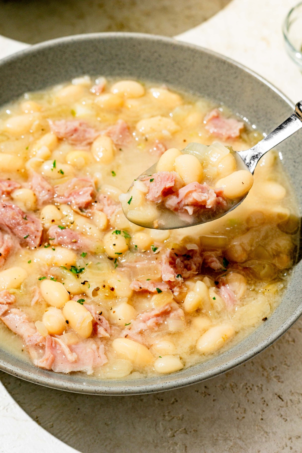 A blue bowl with senate bean soup garnished with fresh parsley. There is a spoon taking a bite of soup from the bowl. The bowl is on a white counter.