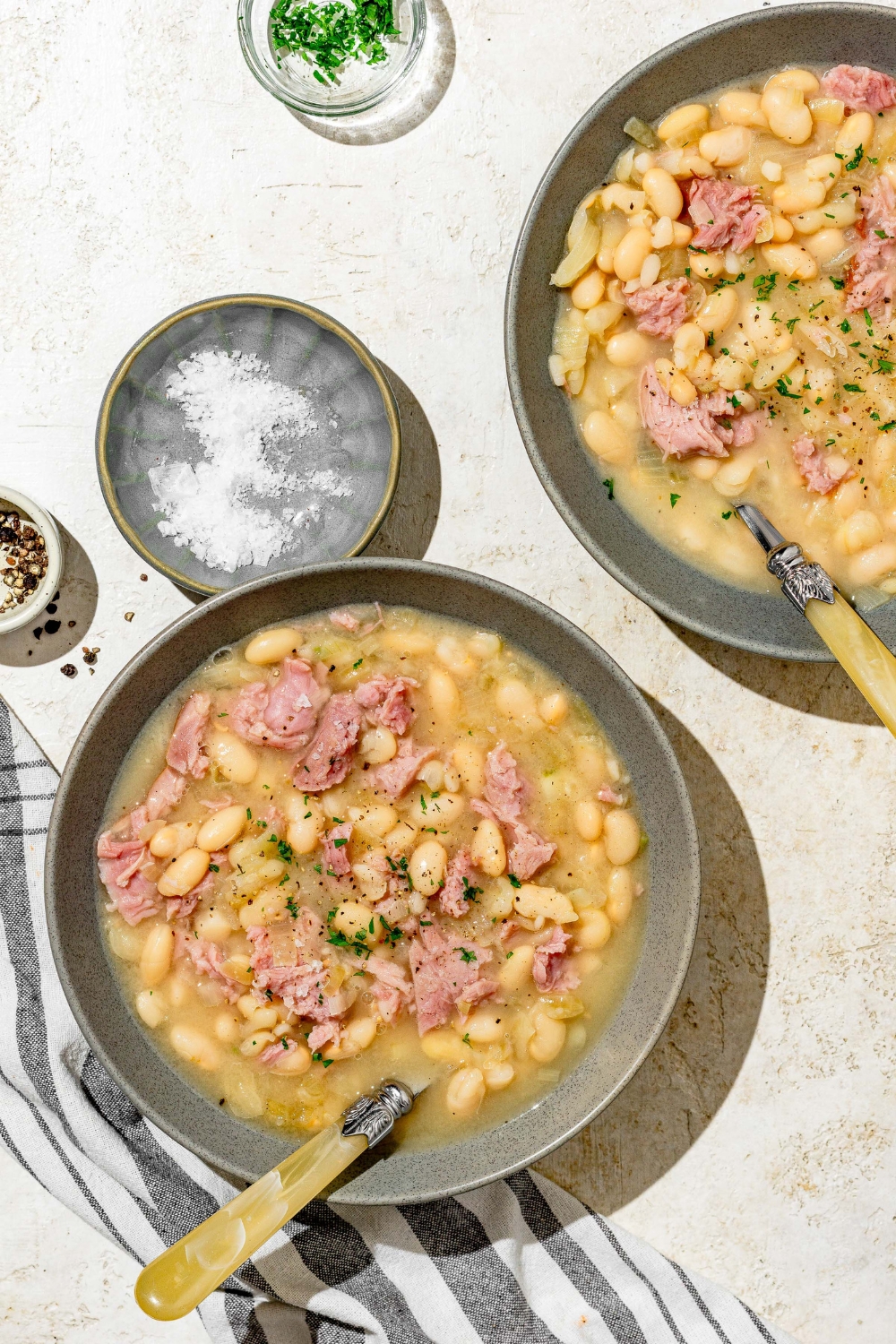 A blue bowl with senate bean soup garnished with fresh parsley. There is a spoon in the bowl. The bowl is on a white counter with an additional bowl of soup and small bowl of garnishes.