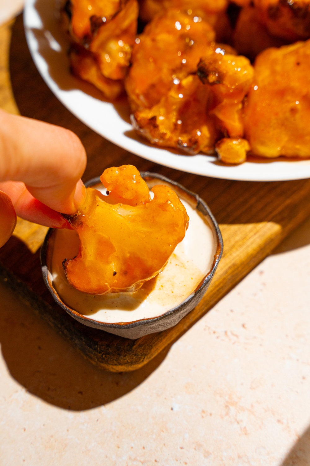 A hand dipping a Buffalo cauliflower wing into a small bowl of dipping sauce. The bowl is on a wooden board with a plate of Buffalo cauliflower wings.
