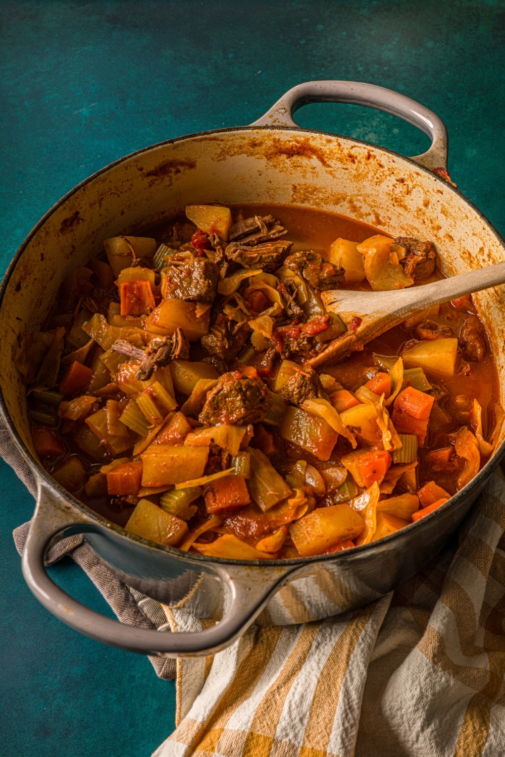 A dutch oven with beef cabbage stew on a blue counter with a striped napkin. A wooden spoon is stirring the stew.