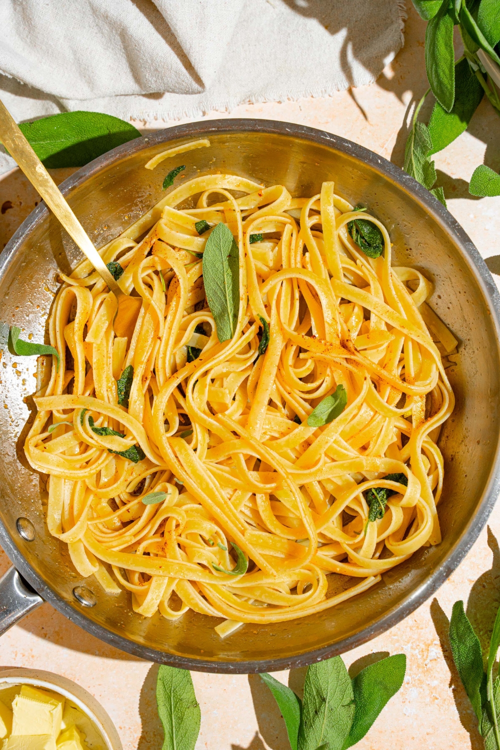 A skillet with a fork twirling a bite of linguine pasta in brown butter sauce with sage. The skillet is on a tan counter with sage and white cloth napkin.