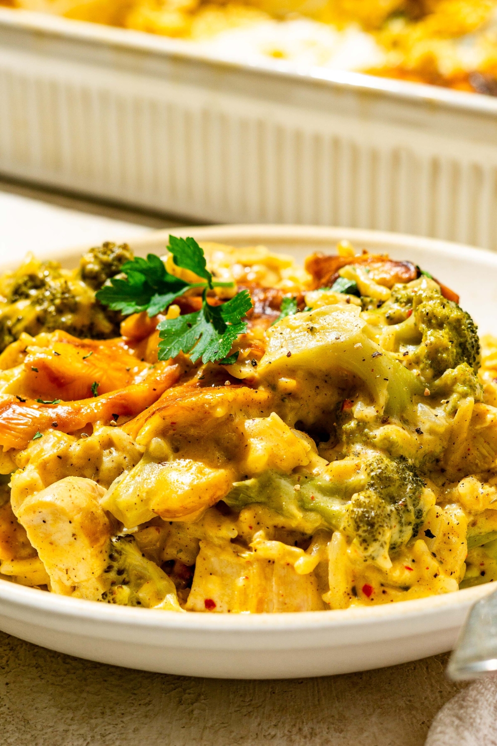 A bowl with chicken broccoli rice casserole garnished with fresh parsley. The bowl is on a tan counter with a baking dish of casserole.