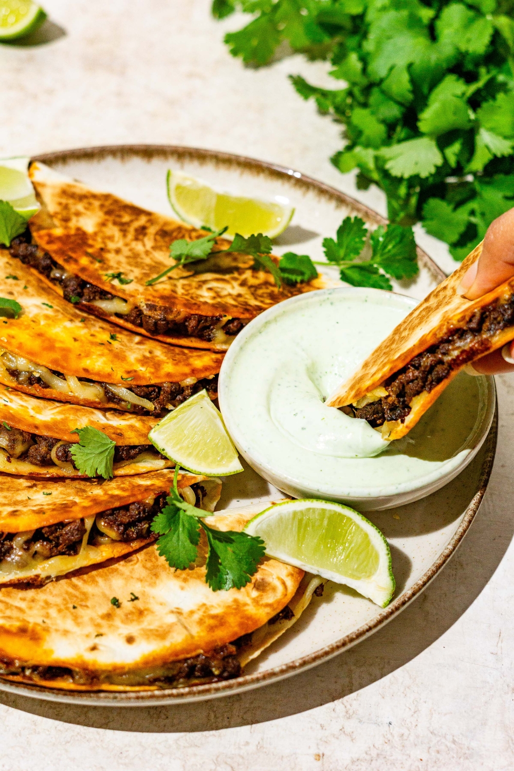 A hand dipping a crispy beef taco into a bowl of sauce. The bowl is on a plate with tacos garnished with cilantro and lime wedges. The plate is on a tan counter with garnishes.