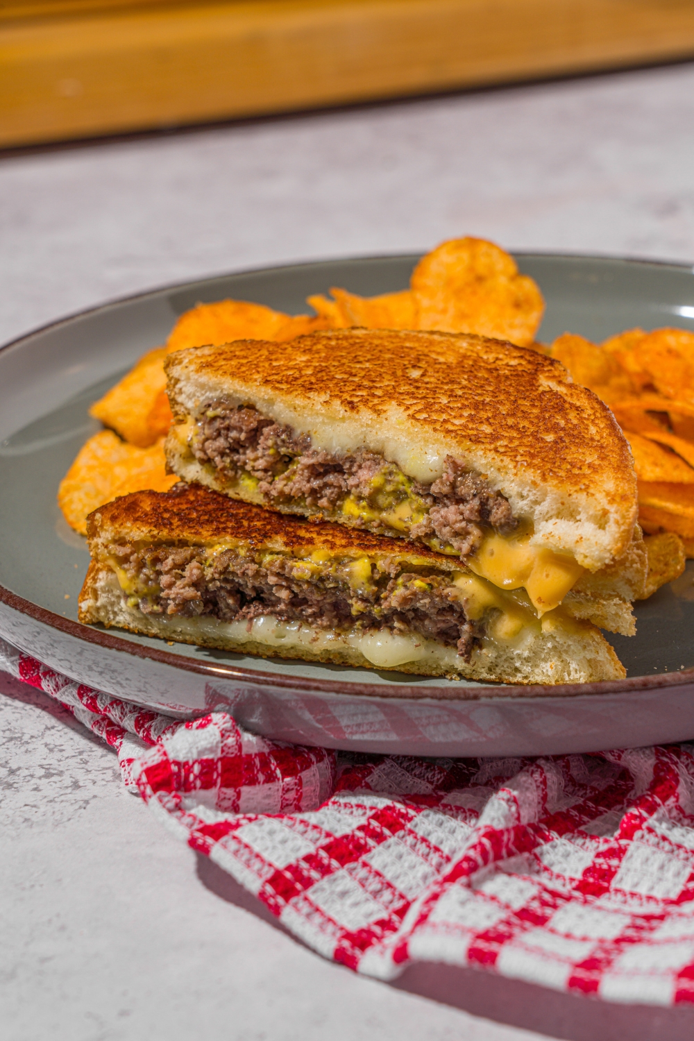 A green plate with a sliced copycat Applebee's grilled cheeseburger stacked on top of each other. There are chips on the plate. The plate is on a stone counter with a red checkered napkin.