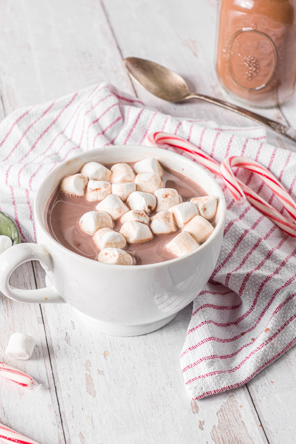 A white mug with homemade hot cocoa topped with marshmallows. The mug is on a wooden counter with a red striped napkin, spoon, and candy canes.