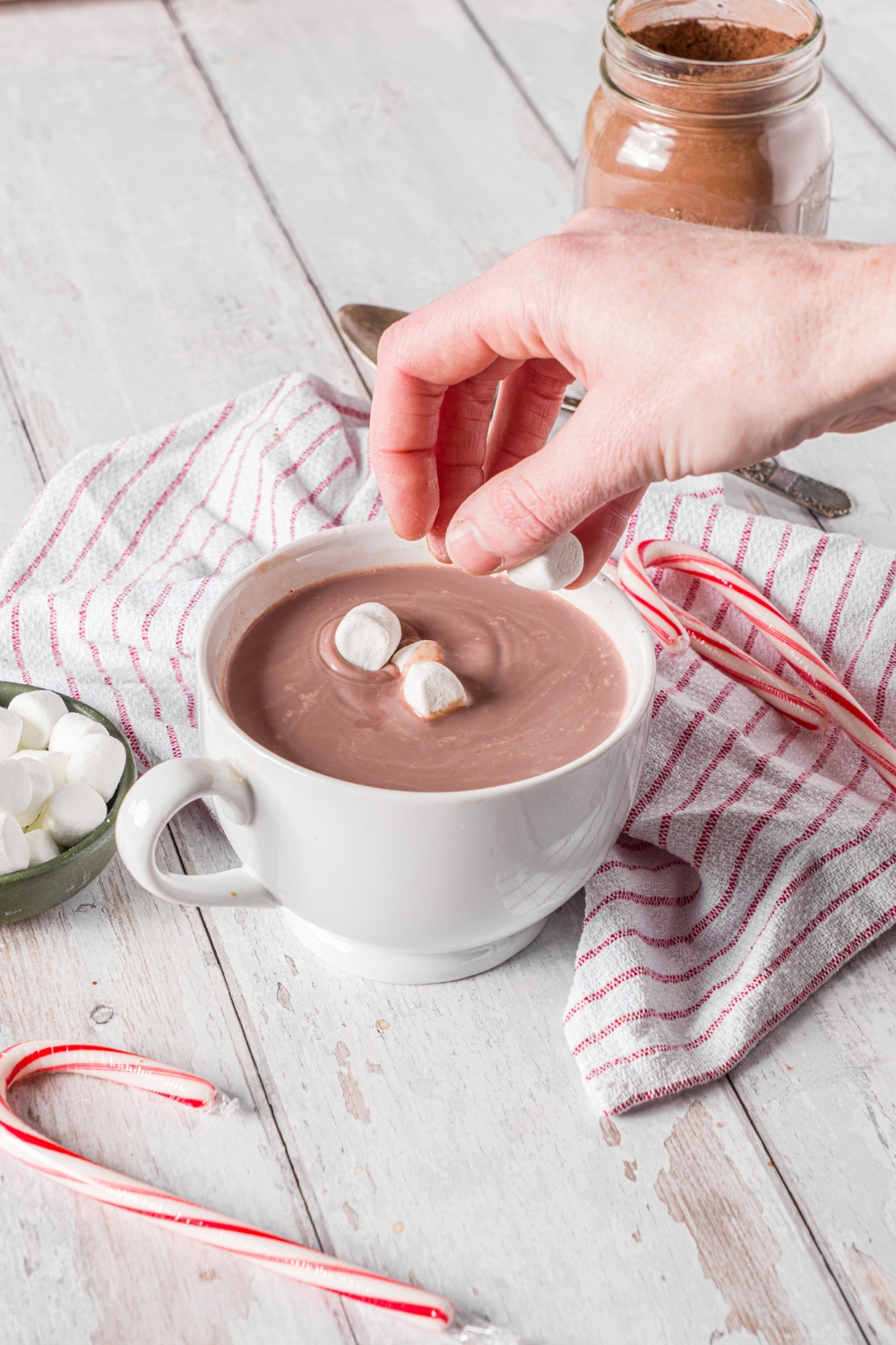 A white mug of homemade hot cocoa with a hand adding marshmallows on top. The mug is on a wooden counter with a small bowl of marshmallows, jar of hot cocoa mix, and candy canes.