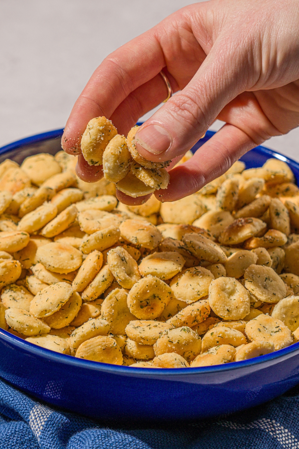 A blue bowl filled with ranch oyster crackers. The bowl is on a white counter with a blue striped napkin. A hand is taking some of the crackers.