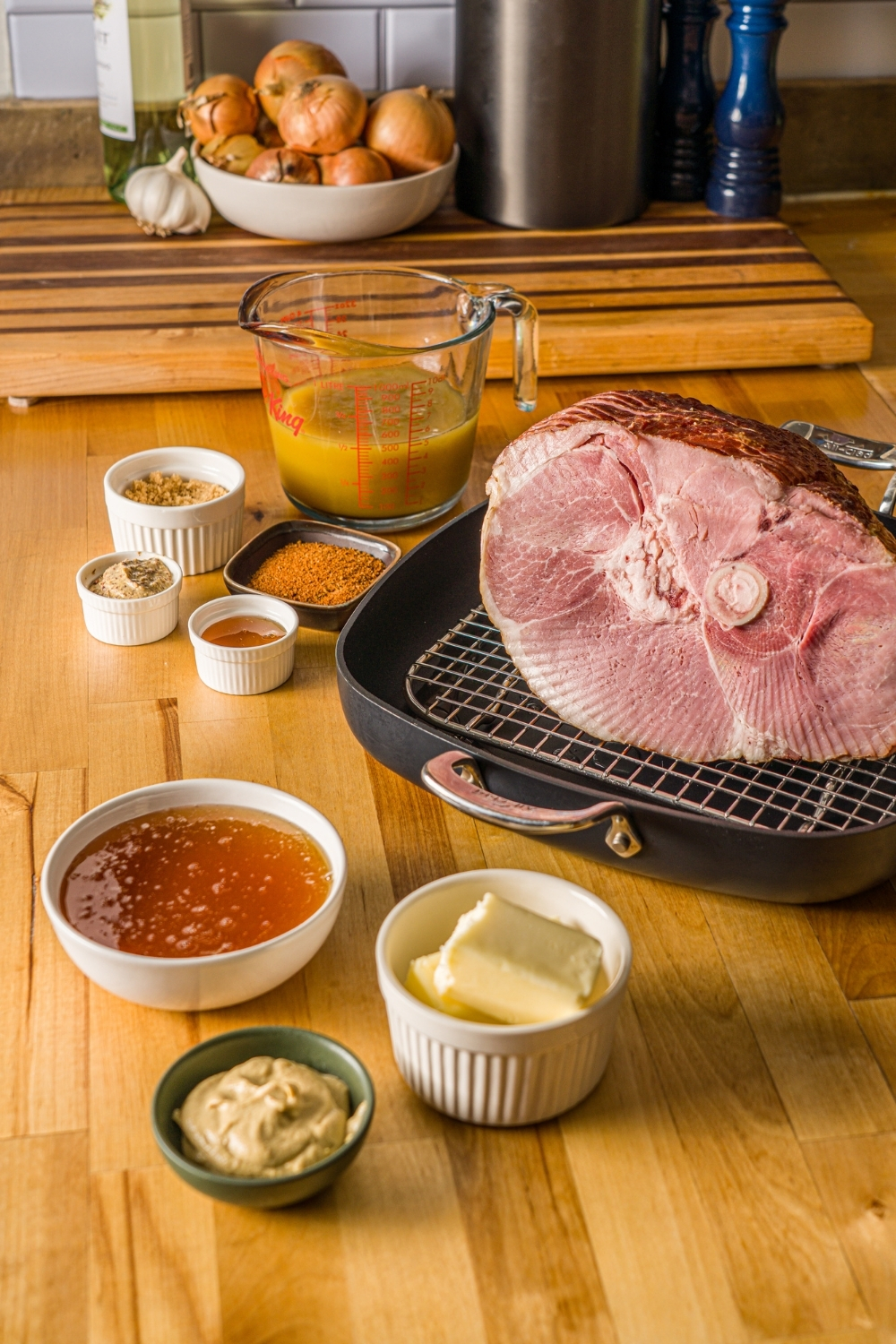 Ingredients to make glazed spiral ham including a spiral ham on a roasting pan lined with a rack, a bowl of honey, butter, mustard, pineapple juice, and seasonings.
