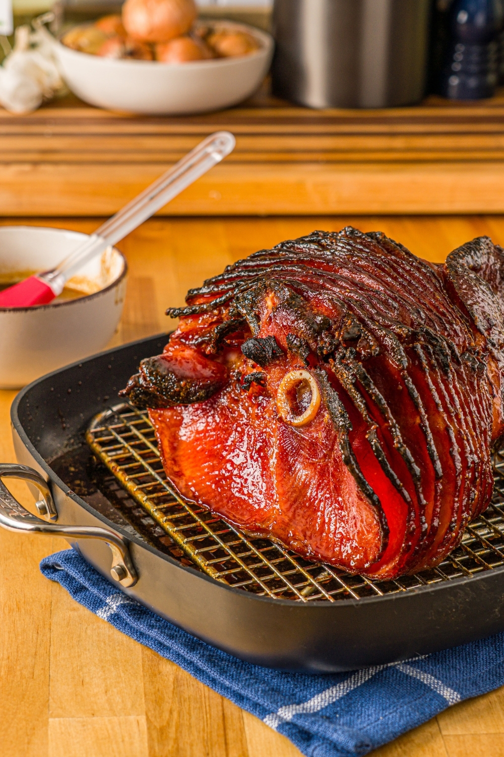 A baked glazed spiral ham in a roasting pan lined with a rack. The pan is on a wooden counter with a blue cloth napkin and small bowl of glaze with a basting brush.