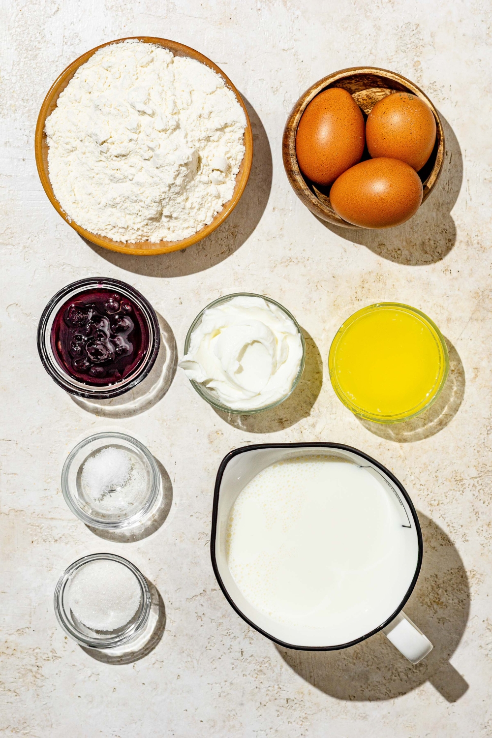 An overhead shot of several bowls in various sizes containing ingredients to make Swedish pancakes including eggs, milk, flour, sugar, butter, whipped cream, and blueberry jam.
