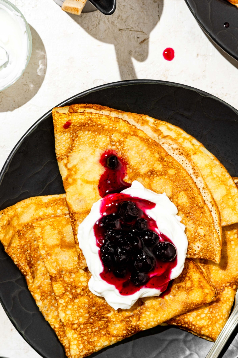 A black palate with Swedish pancakes topped with whipped cream and blueberry jam. The plate is on a white counter.