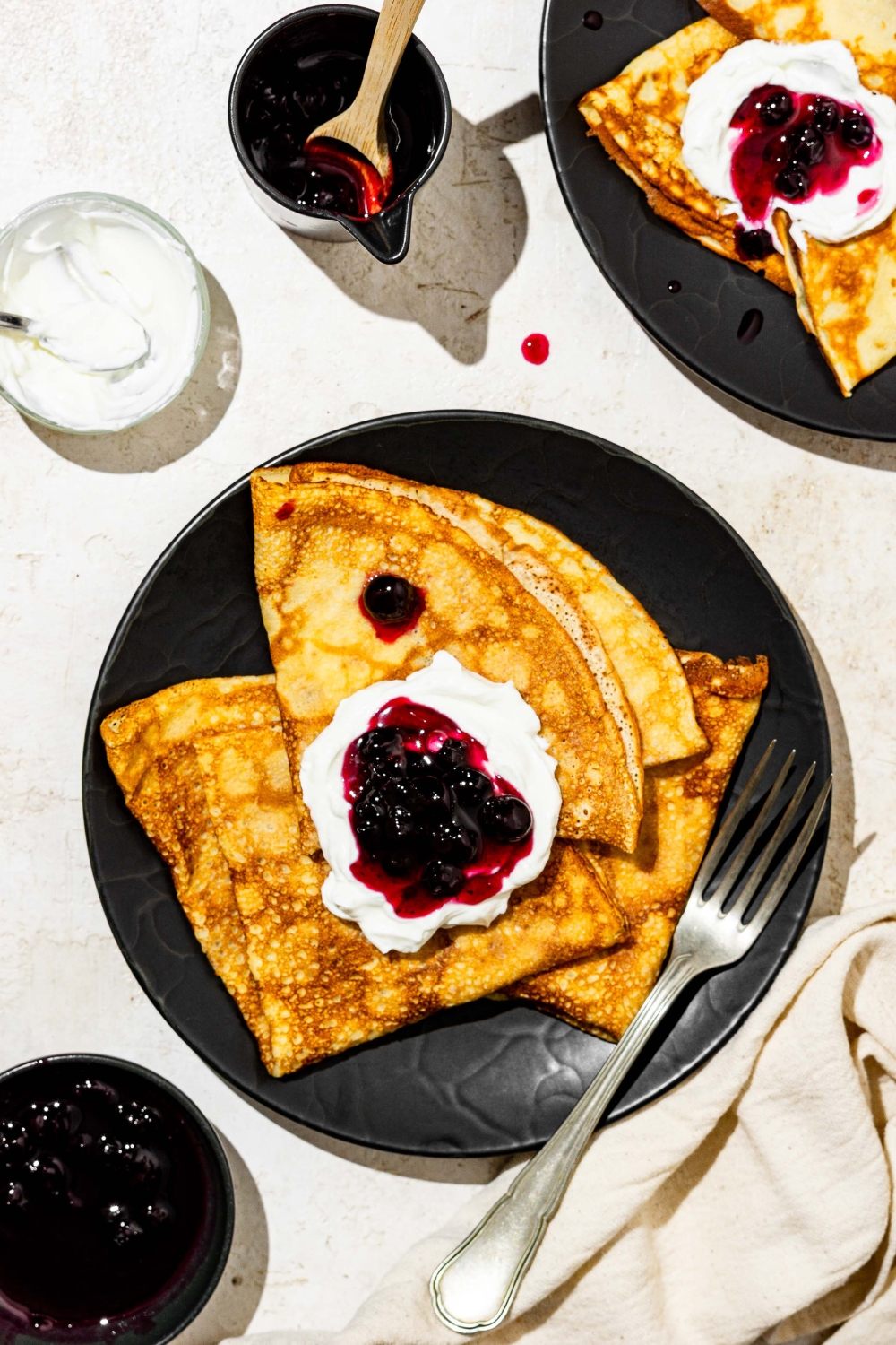 A black palate with Swedish pancakes topped with whipped cream and blueberry jam. There is a fork on the plate. The plate is on a white counter with an additional plate of pancakes and small bowl of jam.