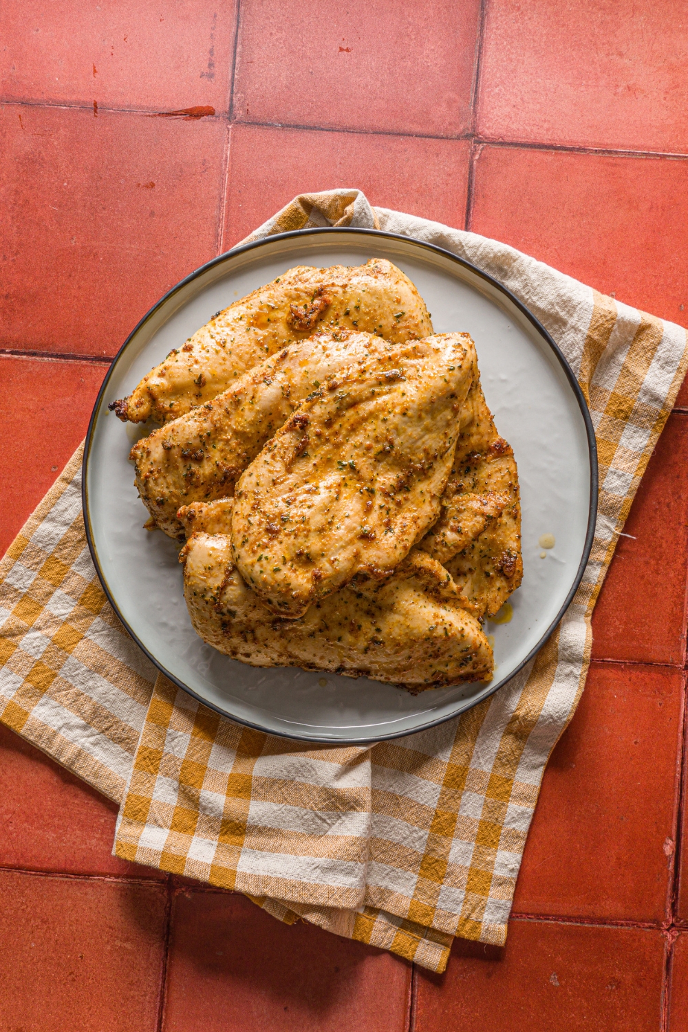 A blue plate with thin sliced oven baked chicken breasts. The chicken is cooked in seasonings and served with lemon wedges. The plate is on a yellow checkered napkin on a red tiled counter.