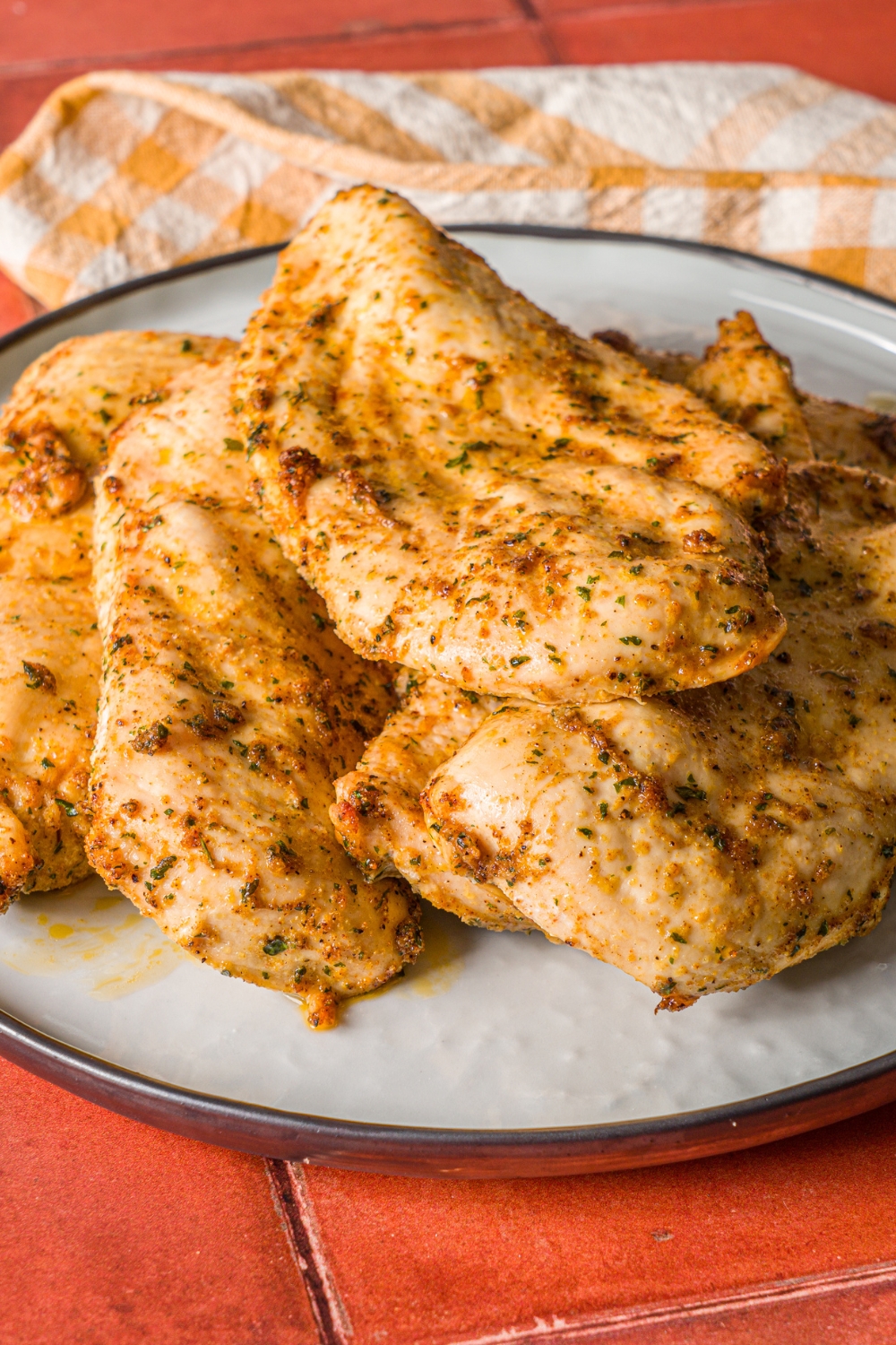 A blue plate with thin sliced oven baked chicken breasts. The chicken is cooked in seasonings and served with lemon wedges. The plate is on a yellow checkered napkin on a red tiled counter.