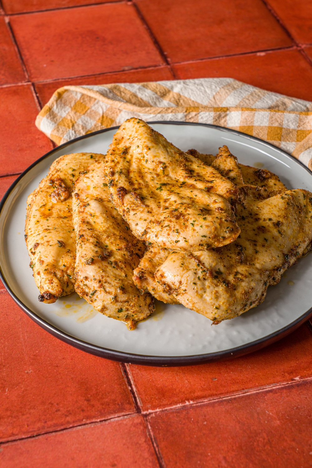 A blue plate with thin sliced oven baked chicken breasts. The chicken is cooked in seasonings. The plate is on a yellow checkered napkin on a red tiled counter.