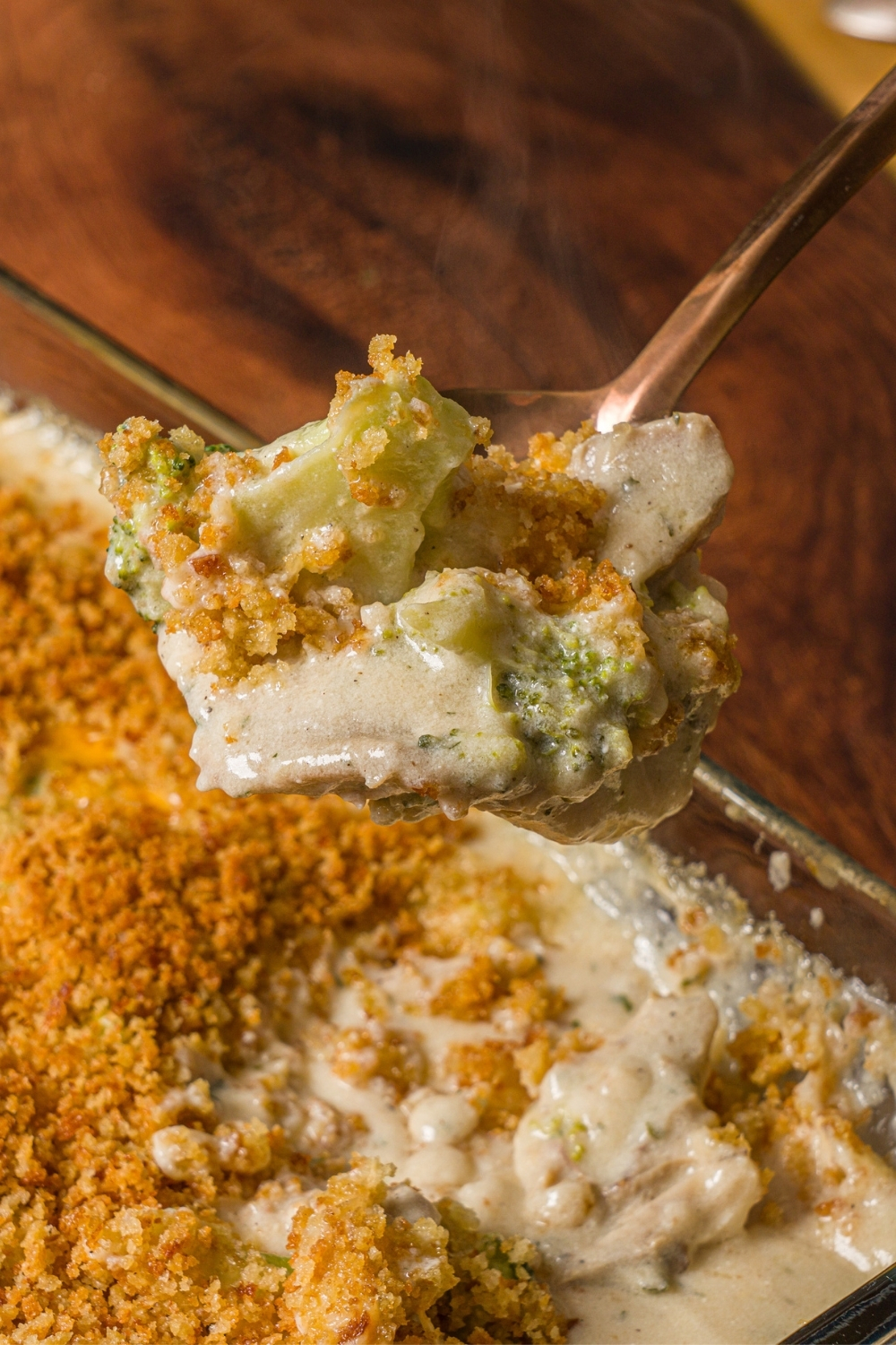 A close up of a fork with a bite of turkey divan. There is a baking dish with turkey divan casserole on a wooden counter in the background.