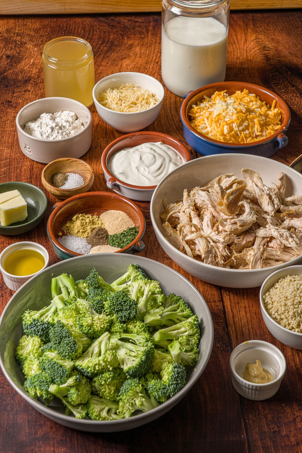 A wooden counter with several bowls in various sizes containing ingredients to make turkey divan including shredded turkey, broccoli, shredded cheese, milk, sour cream, panko breadcrumbs, garlic, flour, butter, oil, and seasonings.