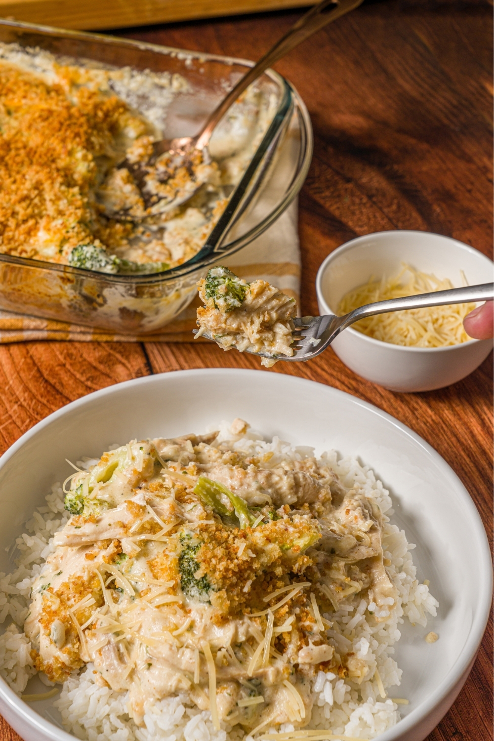 A white plate with white rice topped with turkey divan. A fork with a bite of turkey is over the plate. The plate is on a wooden counter with a baking dish of baked turkey divan and small bowl of shredded cheese.