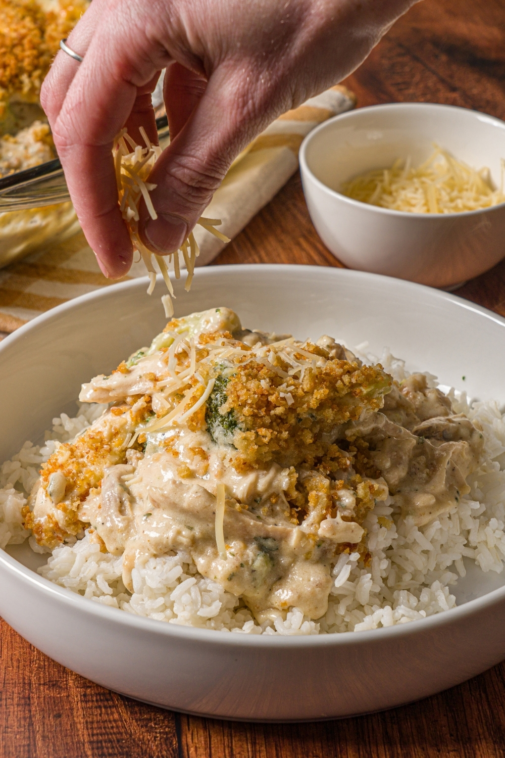 A white bowl with white rice topped with turkey divan. A hand is sprinkling shredded cheese over the bowl. The bowl is on a wooden counter with a small bowl of shredded cheese.