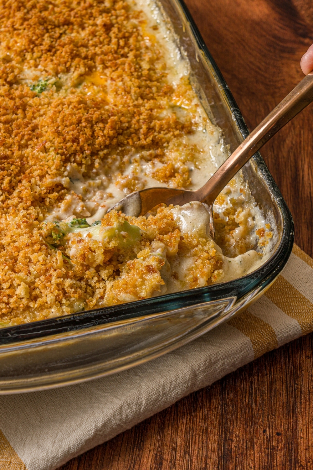 A baking dish with baked turkey divan topped with panko breadcrumbs. A spoon is scooping a bite of turkey from the dish. The dish is on a wooden counter.