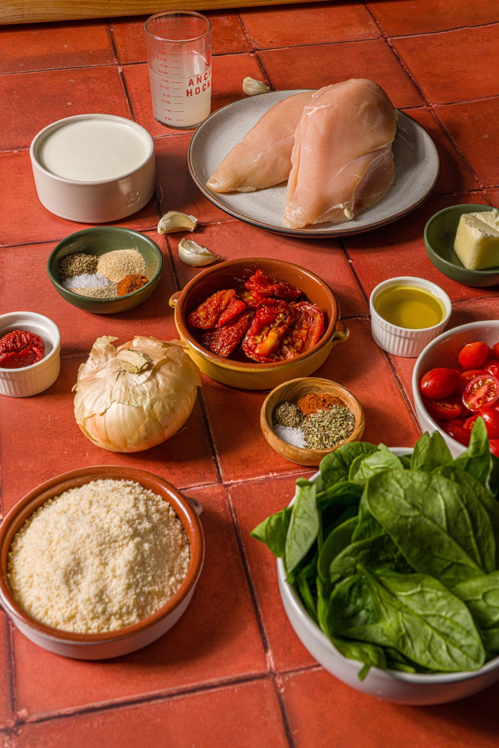A tiled counter with ingredients to make tuscan chicken pasta including uncooked chicken breasts, spinach, cherry tomatoes, garli, onion, tomato paste, sundried tomatoes, heavy cream, and seasonings.