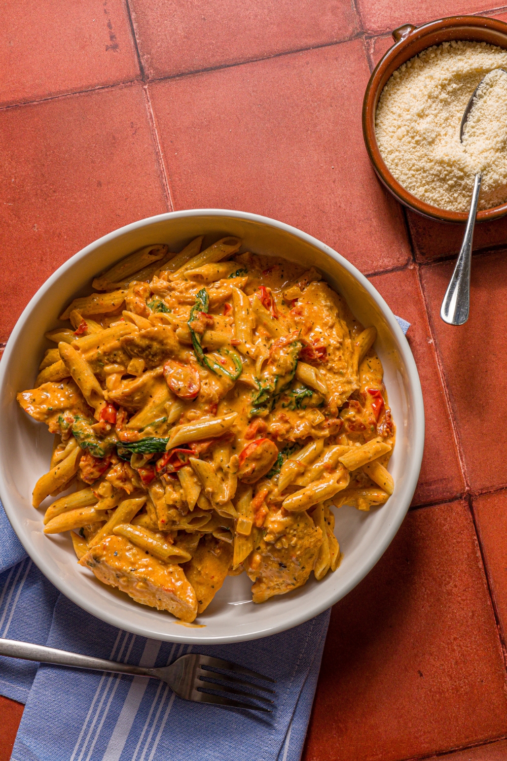 A white bowl with tuscan chicken pasta on a tiled counter with a blue cloth napkin and small bowl of parmesan cheese with a spoon in it.