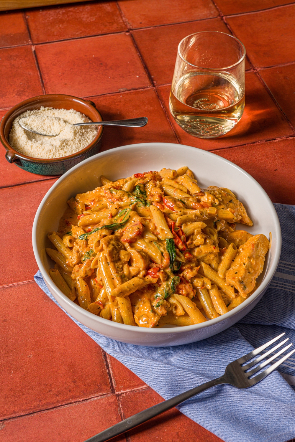 A white bowl with tuscan chicken pasta with a spoon of parmesan sprinkling over the bowl. The bowl is on a tiled counter with a blue cloth napkin, glass of white wine, and small bowl of parmesan.