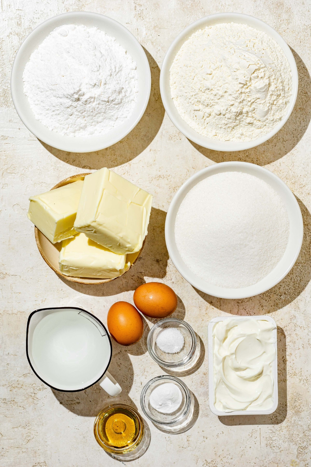 An overhead shot of several bowls in various sizes containing ingredients to make white Texas sheet cake including flour, sugar, butter, eggs, vanilla, baking powder, sour cream, and powdered sugar.