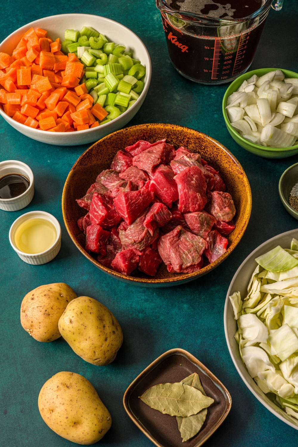 Several bowls in various sizes containing ingredients to make beef cabbage stew including cubed beef, potatoes, onions, cabbage, carrots, celery, and beef stock.