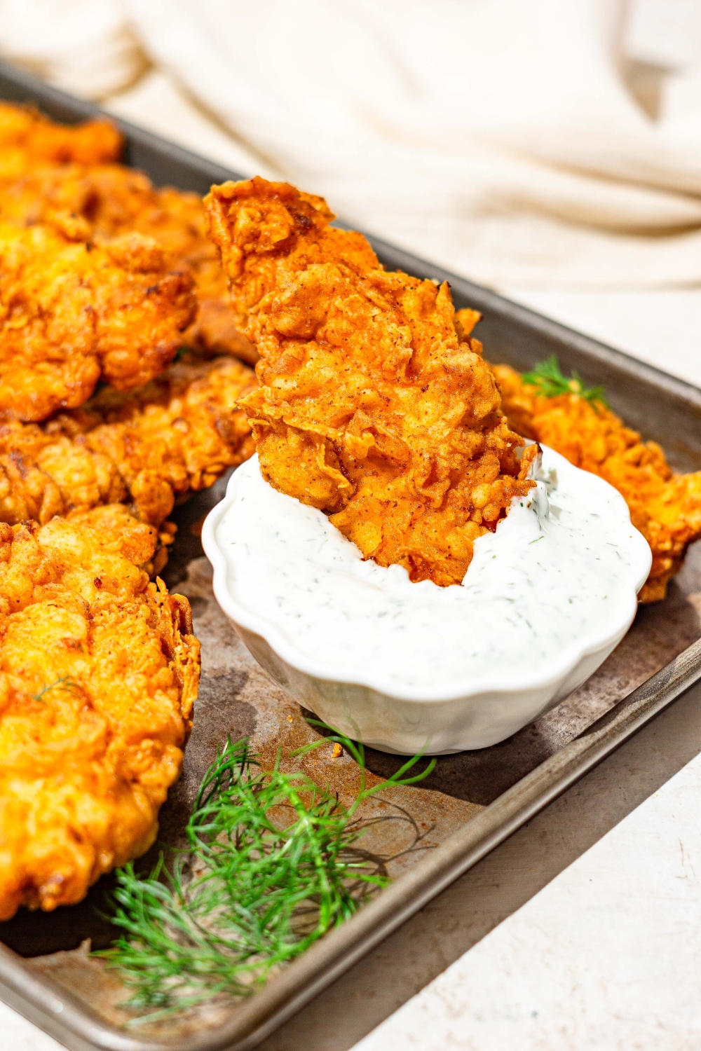 A baking sheet with buttermilk fried chicken tenders with a bowl of homemade ranch dressing. A tender is dipped in dressing. The sheet is on a white counter with a white cloth napkin.