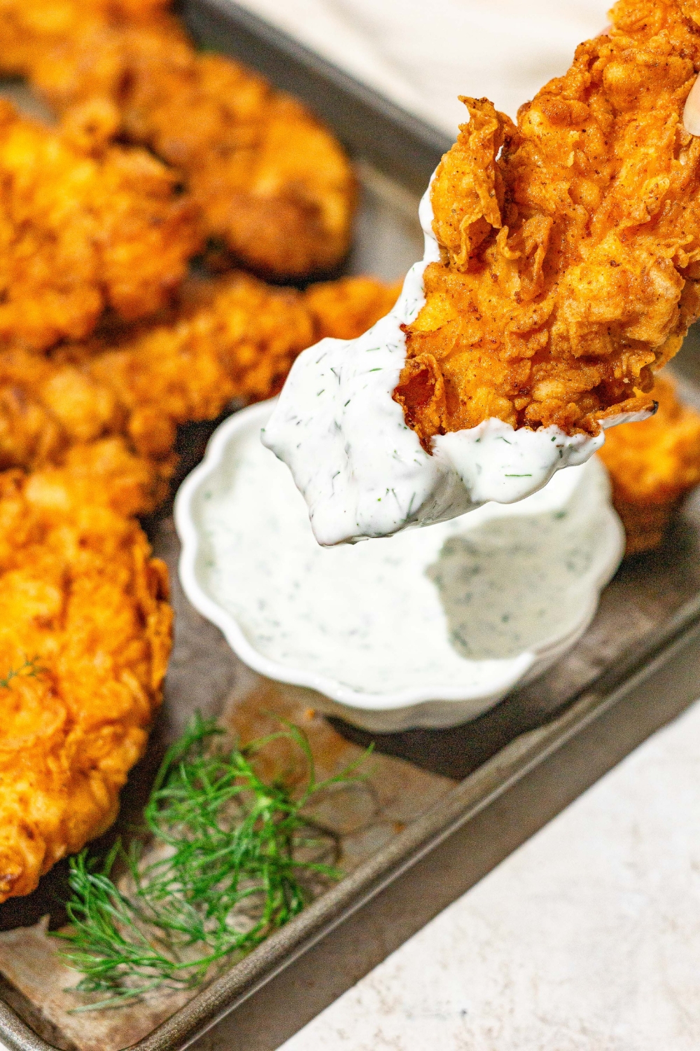 A baking sheet with buttermilk fried chicken tenders with a bowl of homemade ranch dressing. A tender is dipped in dressing. The sheet is on a white counter and garnished with a sprig of dill.